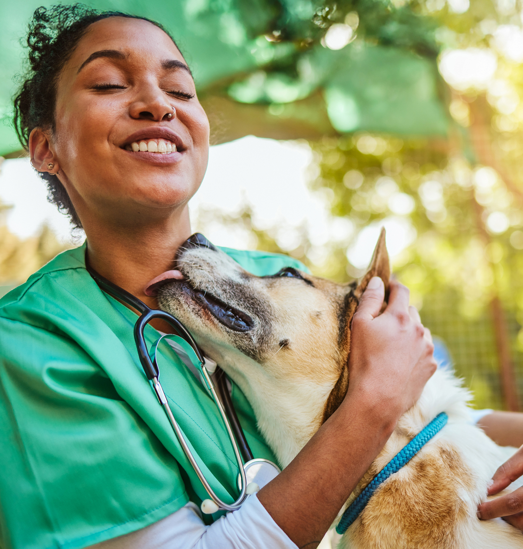A smiling veterinarian in green scrubs hugging a happy dog.