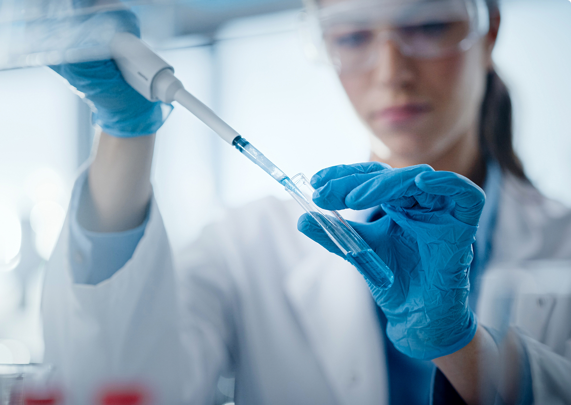 A female scientist in a lab coat and gloves using a pipette to transfer a blue liquid into a test tube.