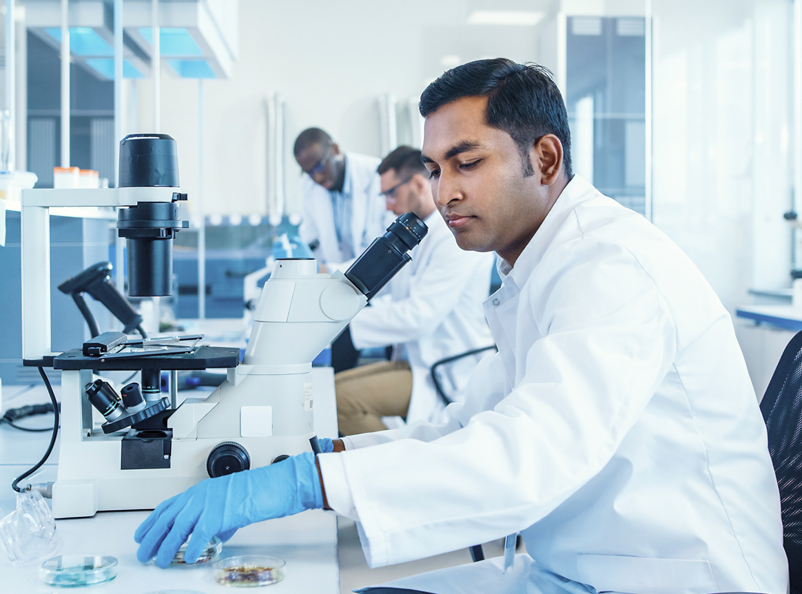 A male scientist in a white lab coat using a microscope in a laboratory with other scientists working in the background.